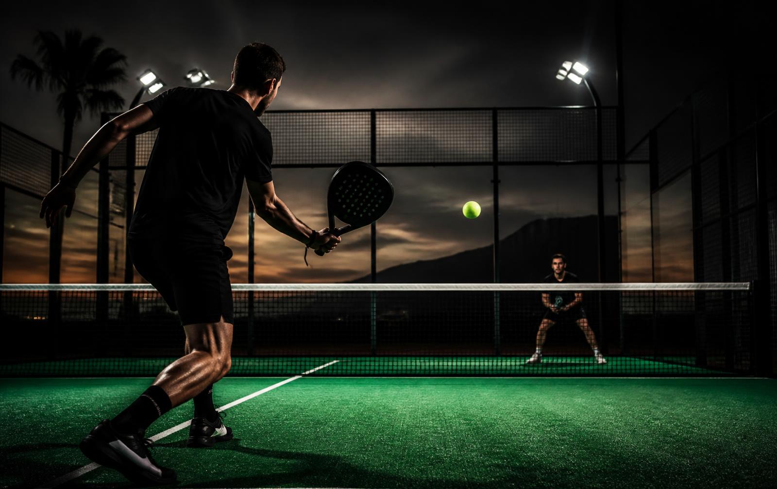 Two players mid-rally on a glass padel court at dusk